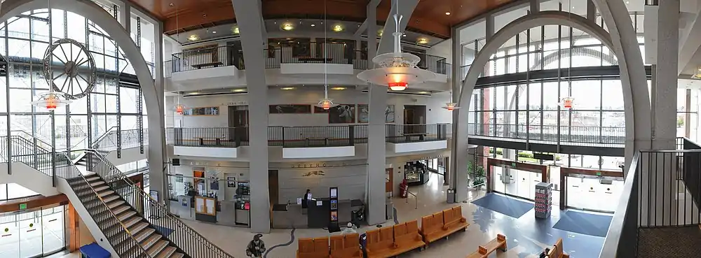 The lobby of a large building, with balconies from two upper stories framed by large steel arches.