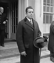 A black and white portrait of a formally dressed man standing outside a building. He is wearing a dark coat, white shirt, dark tie and he is holding a hat with his left hand.