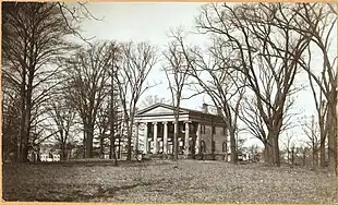 Photograph of a large house atop a shallow slope.  The roof is supported by a row of ionic columns.  Many large trees surround the house.