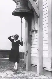 Photo of keeper Fannie Salter standing next to the Turkey Point Light fog house. She is looking out to sea and holding the rope to ring the large fog bell at the station.