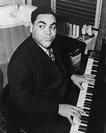 Stocky African-American man sitting and playing the piano. He has black hair and thick black eyebrows, and is grinning and looking to the left. The man is wearing a striped black suit, white shirt and a tie.