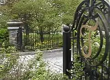 An open gate revealing the road to enter a cemetery, surrounded by grass, flowers and trees.