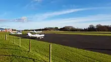 A tarmac runway with a small plane landing is shown in the background. Small planes are also parked in the forecourt