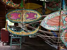 A wau-maker's workshop. Foreground: Wau frames. Background: A completed wau. Further background: The wau-maker.