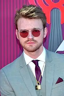 An image of a blonde-haired man wearing a suit and sunglasses against a purple-red backdrop.