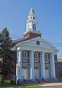 A two-story brick building with a white colonnade on front, topped by a green rounded cupola, in the American Colonial Revival style. "First Congregational Church" is engraved across the front above the columns. It is seen from across the street, with wires in front of it.