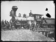 Steam locomotive partly on small railroad bridge; several men standing on and around it