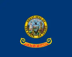 A blue flag with a circular seal in the center. The words "State of Idaho" appear in gold letters on a red and gold band below the seal.