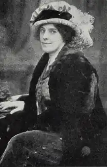 A white woman, seated, wearing a large plumed hat, and a dark jacket, possibly velvet; her skirt has a beaded or button detail down the side seam