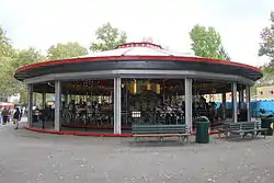 A small circular building with a saucer-like red and gray roof, on an asphalt surface, with trees behind it