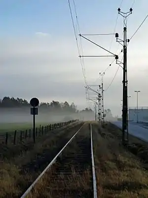 A railway line with overhead wires, supported by latticework gantries on its right, seen just from its side. On the left is the rear of a sign with a circular piece above an equally-sized rectangular one. Golden-colored grasses rise from in between the ties, and there is a fogged area of woodland around the tracks in the distance under clouds with blue sky above them. On the right is a road; a fence separates the track right-of-way from a green field on the left.