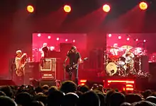 A crowd of people standing before a stage lit by four lights from above. On the stage, from left to right, is a man with a guitar, a man dressed in black holding a guitar, and a man sitting behind a drum set.