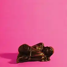 A photo of a bronze statue of a nude woman, tied up and lying on the ground in the fetal position, over a pink background.