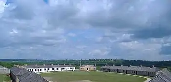 A photograph of a set of long, single-story buildings arranged in a circle around a small grassy field.