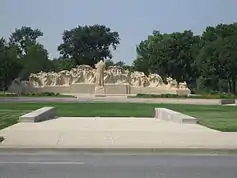 A large sculpture of many figures opposed by a single figure with large trees in the background.