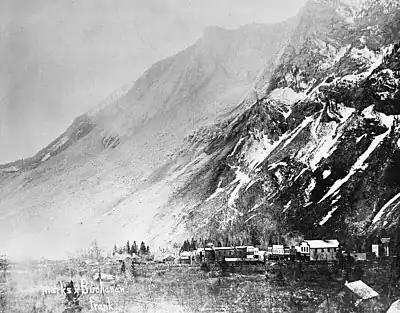 A small town shown at the base of a mountain. The mountain's face stands barren following a large rockslide and a light cloud of dust is visible in the air.