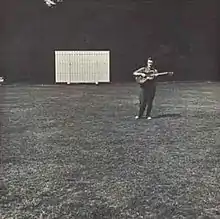 The album cover is a black-and-white photograph of a sports field with a score board in the distance, off-center to the left. Off-center to the right and also in the distance Fred Frith is standing playing a guitar. There is no text on the cover.