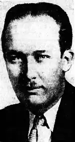 Black-and-white photographic portrait of an expressionless adult man with slicked-back hair, a toothbrush mustache, and a poorly defined cheekbone. His head appears bulbous, being bloated around the forehead and tapering toward the chin. One eye is less well defined. He is depicted wearing a suit.