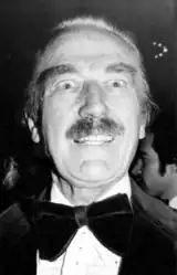 Black-and-white portrait photograph of a wide-eyed man with large, suspended eyebrows and a full mustache. His mouth hangs agape, allowing his perfect teeth to flash through. He wears a velvety tuxedo with a considerably large bowtie. The crescent of a receded hairline is backlit, and passersby populate the otherwise dark background.