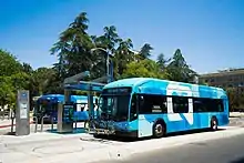 A teal blue bus stopped at a station, with another bus in the distance