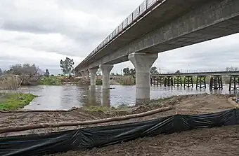 A nearly completed concrete bridge crosses a river filled with water on a cloudy day