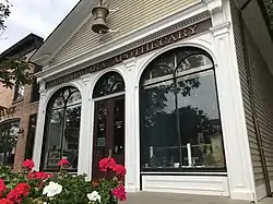 A cloudy sky is visible in the top left, behind the gable of a roof at the front of which is mounted a large, bronze, ornamental mortar and pestle over a clapboard siding. Below the gable is a narrow masthead reading "Niagara Apothecary" in gold lettering contrasted by a dark red background. Below that are two large arch windows flanking a French door. To the left is a portion of a brick building, and at bottom left are flowers.