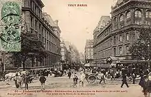 A sepia photograph looking down a built-up street, with the tramway to the right. A small crowd waits to board the tram.