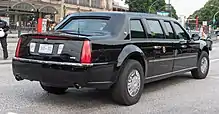 A black Cadillac limousine is at a 45-degree angle, showing to the camera its rear and starboard sides.  The trunk sports two antennae, there is a presidential seal on the rear passenger door, and the license plate is numbered "800 002".