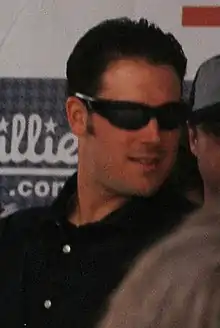 A dark-haired man with sideburns wearing a white shirt and dark sunglasses