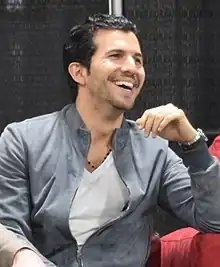 A man with black hair laughing while facing to the right of the camera.