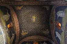 Interior image of the relatively small sail vault ceiling at the cross intersection of the Mausoleum of Galla Placidia with intact mosaic decoration of gold stars on a blue background and a gold cross at the center