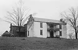 A two-story white house with a porch and a balcony over it, flanked by two trees. A car from 1930s is parked at the house.