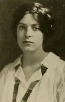 A young white woman with dark hair loosely braided across the crown, wearing an open-collared white blouse.