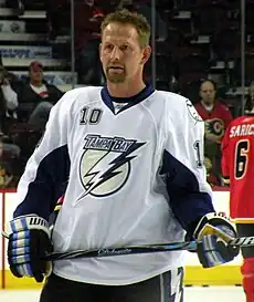 Upper body of a man with short, brown hair and a goatee looks into the distance. He is in a full hockey uniform, without helmet; the jersey is white with blue trim and has a stylized lighting bolt logo with the words "Tampa Bay"