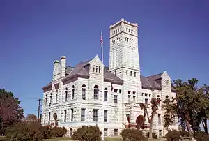 A rough-faced light-colored stone building with pointed roofs and a tower in the center amidst trees and shrubs. An American flag flies from a white pole in the front