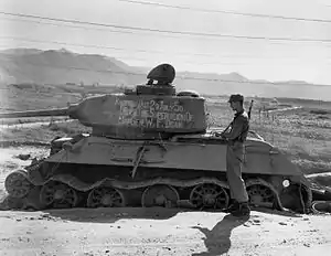 Soldiers stand around a destroyed tank with writing on it reading "Knocked out 20 July 1950 with the supervision of Maj. Gen. W.F. Dean"