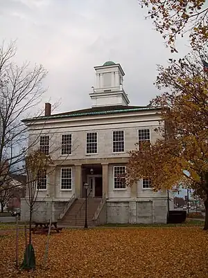 A two-story gray stone building on a raised basement with entry steps leading to the front door and a wooden cupola rising from the center of the roof photographed on a cloudy day with autumn leaves on a surrounding tree and the ground in front.