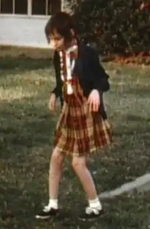 A slightly blurry color picture of Genie, facing slightly right of the camera, walking by herself outside in the hospital recreation yard. She is wearing a plaid-patterned dress and thin sweater and looks extremely pale, emaciated, and expressionless. Her limbs are exposed and look extremely thin. Both of her knees are very bent, and her arms are bent forward with both hands hanging down as she holds them out in front of her.