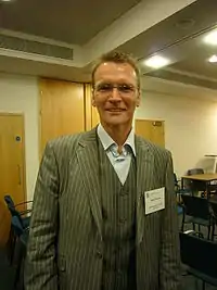 Head-and-torso photograph of Geoff Thomas standing in a lecture room wearing a grey chalk-stripe suit and waistcoat, and an open collared blue shirt