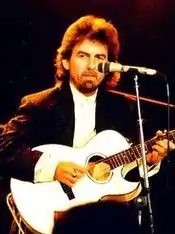 A man with long dark hair and beard, playing a white guitar