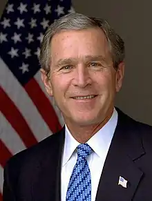 Head and shoulders shot of a man in a dark suit, white shirt and striped blue tie. A United States flag can be seen behind him.