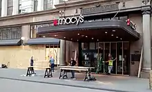 carpenters in front of Macy's Herald Square entrance cutting lumber on sawhorses, erecting wooden frames in front of doors and windows, and affixing plywood to them.