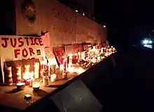 George Floyd–related and Black Lives Matter–themed protest signs and lit candles placed on the base of the Astoria Park War Memorial; the visible portion of the memorial's inscription reads:"GREATER LOVE HATH NO MAN THAN THIS THATA MAN LAY DOWN HIS LIFE FOR HIS FRIENDSERECTED 1926 BY THE PEOPLE OF LONG ISLAND CITY"In the background the glaring headlights of a police cruiser.