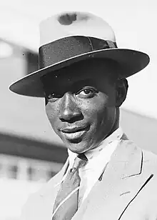 A head and shoulders black and white photograph man in a broad-brimmed hat and suit smiling at the camera.