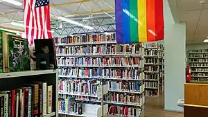 Partial view of the library stacks, with six rows of stacks of bookshelves. Part of a rainbow flag and United States flag are visible hanging from the ceiling.