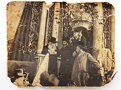 A sepia photograph shows a rabbi standing on the synagogue podium, addressing the congregation. He is reading from a sheet of paper, and three other men stand to his left. Behind him can be seen the white columns and ornate carvings of the holy ark. A chandelier, suspended from the ceiling, appears at the top right.