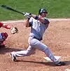 A man in a grey baseball uniform and navy blue batting helmet swings through a pitch.