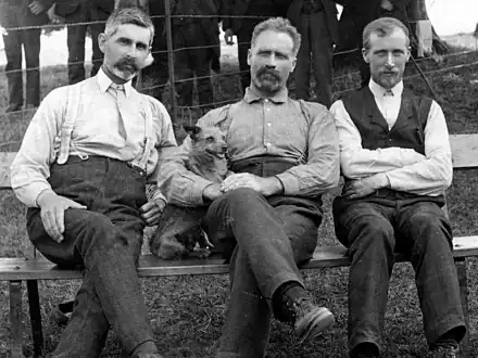 Old photograph depicting three bearded men seated outdoors on a bench with the man in the center holding a small dog