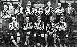 A group of men pose in two lines, one standing and one seated.  Eleven of the men are wearing striped football shirts, long shorts and socks with shinpads worn over them.  Of the other men, one is wearing a suit and tie and a flat cap, and the other is wearing a blazer, waistcoat, shirt without a tie, and a cricket-style cap.