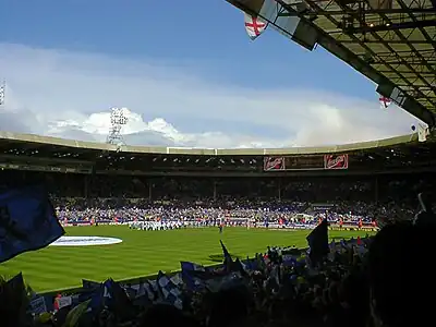 A sports stadium full of spectators.  Those nearest the camera are waving blue and white flags.